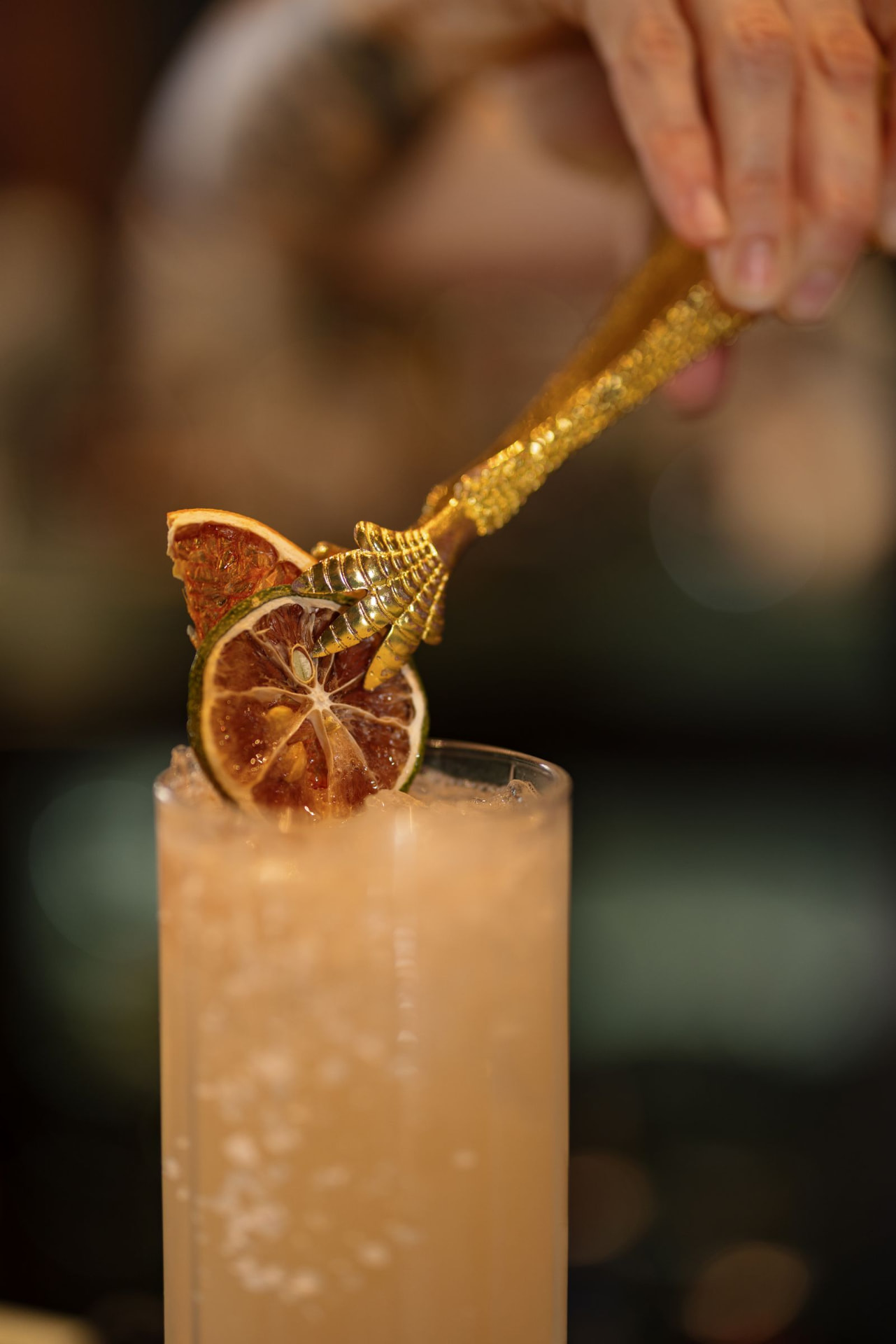A gold claw placing a dried citrus garnish on a highball cocktail