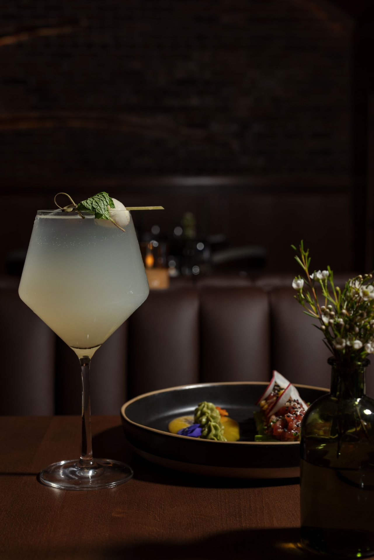 A pale cocktail, plated dish, and bud vase against a dark lounge booth
