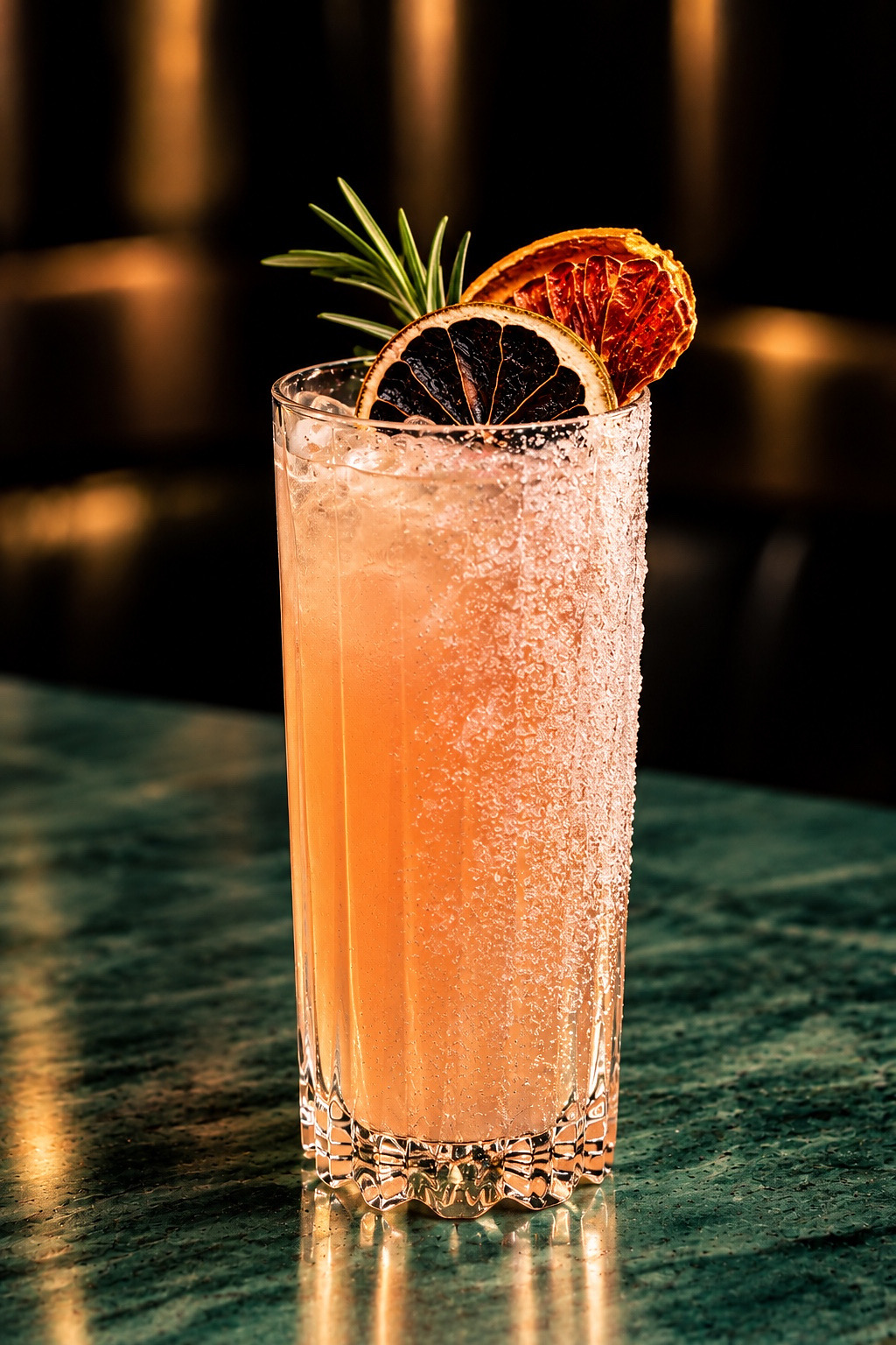 A tall salted-rim cocktail garnished with dried citrus and rosemary on green marble